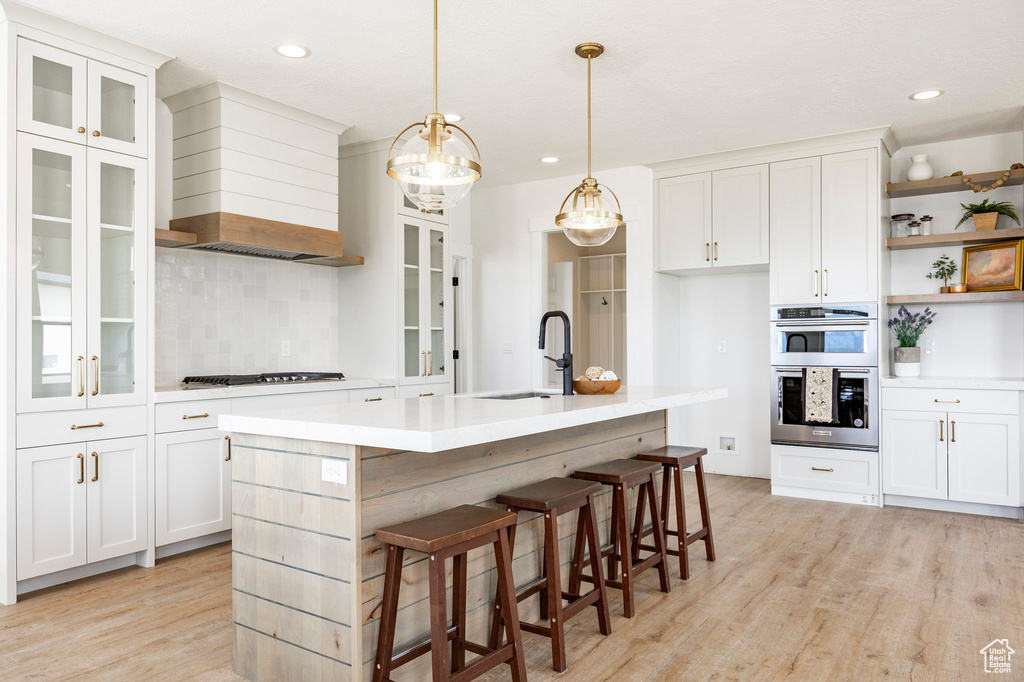 Kitchen featuring custom exhaust hood, open shelves, light wood-type flooring, appliances with stainless steel finishes, and decorative backsplash