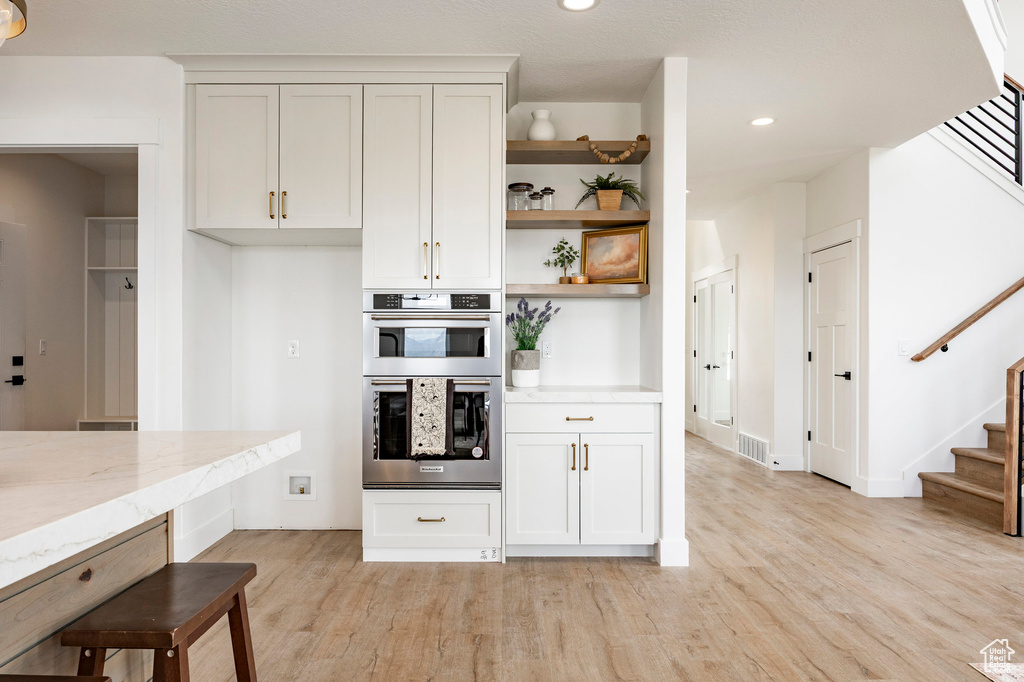 Kitchen with open shelves, light wood-type flooring, double oven, light stone countertops, and recessed lighting