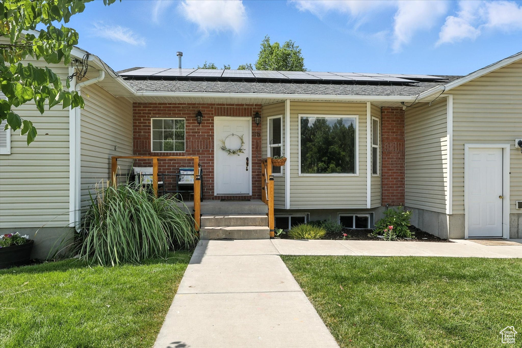 View of exterior entry with roof mounted solar panels, a lawn, brick siding, and roof with shingles