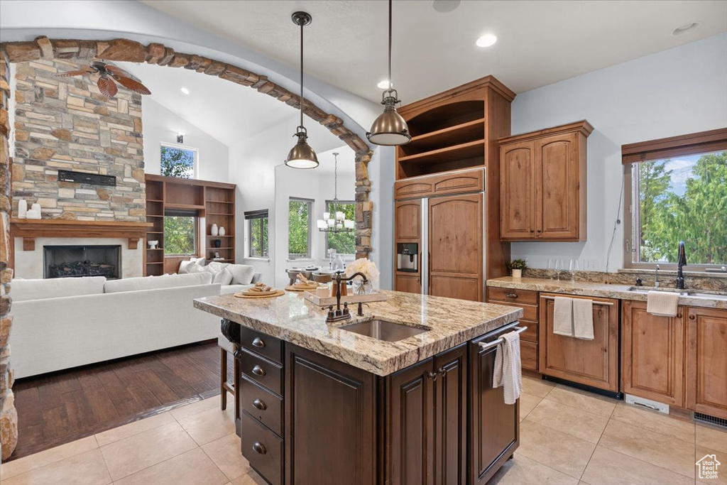 Kitchen with light tile patterned floors, paneled appliances, a center island with sink, light stone countertops, and hanging light fixtures