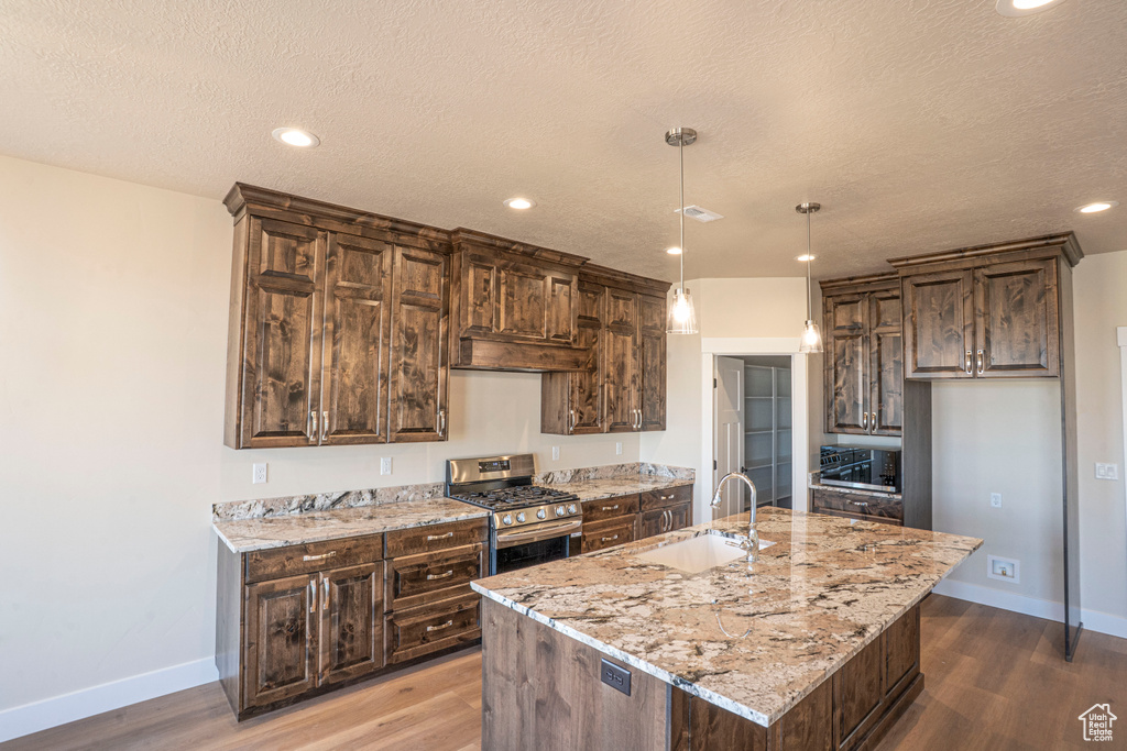 Kitchen with stainless steel gas range oven, wood finished floors, a kitchen island with sink, dark brown cabinetry, and recessed lighting