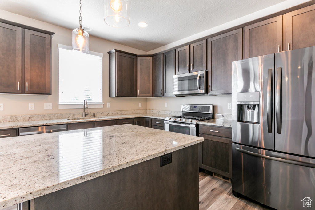 Kitchen featuring stainless steel appliances, dark brown cabinetry, light wood-style floors, light stone countertops, and decorative light fixtures