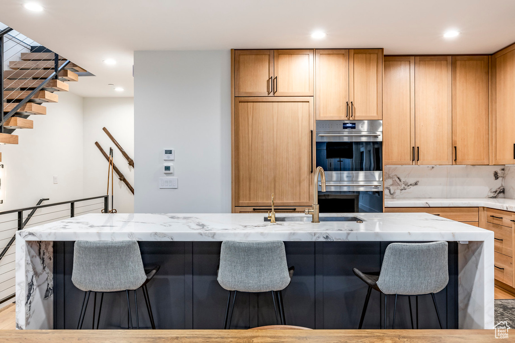 Kitchen featuring stainless steel double oven, light stone counters, a kitchen breakfast bar, recessed lighting, and a center island with sink