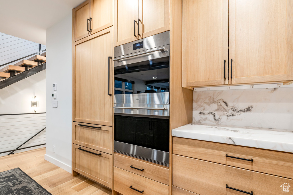 Kitchen with light brown cabinets, double oven, light wood-type flooring, light stone counters, and tasteful backsplash