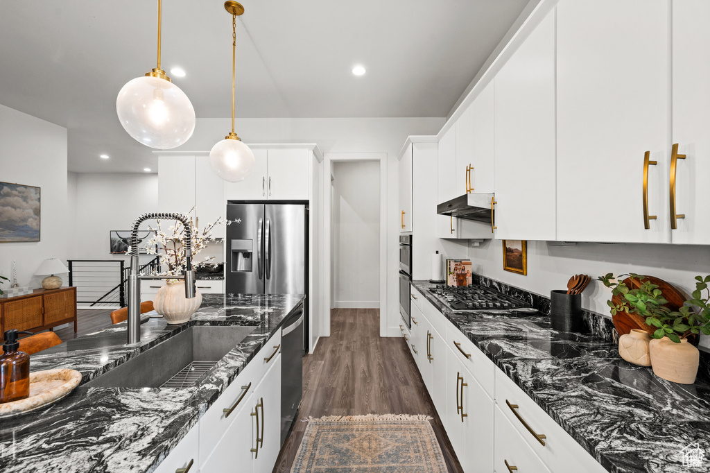 Kitchen with under cabinet range hood, stainless steel appliances, dark stone counters, hanging light fixtures, and white cabinetry