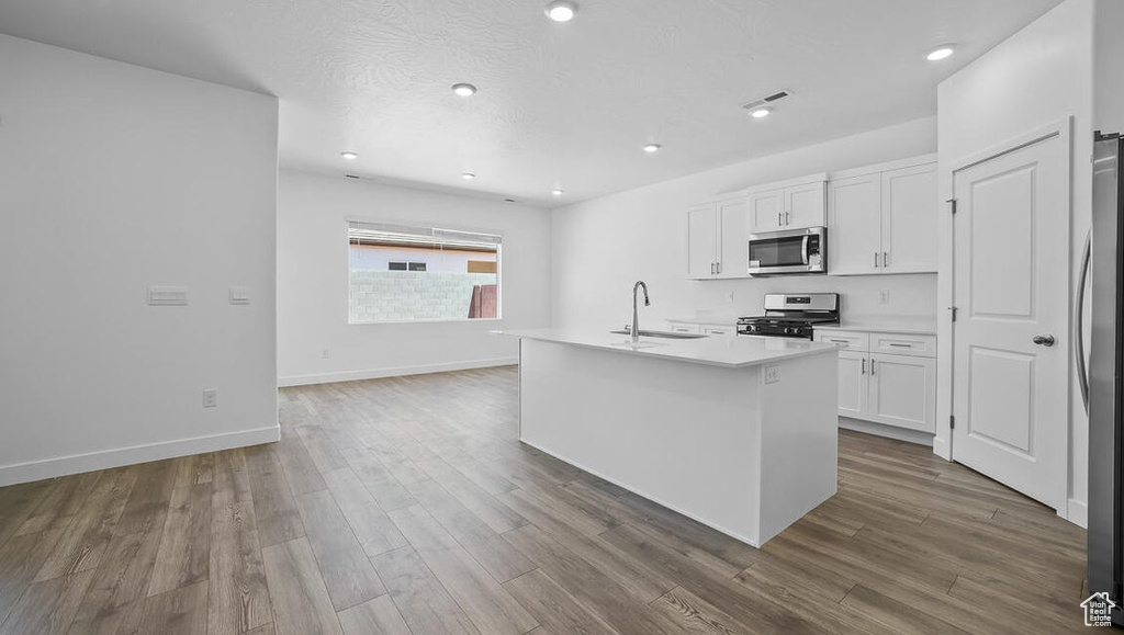 Kitchen featuring appliances with stainless steel finishes, white cabinetry, wood finished floors, a center island with sink, and light countertops