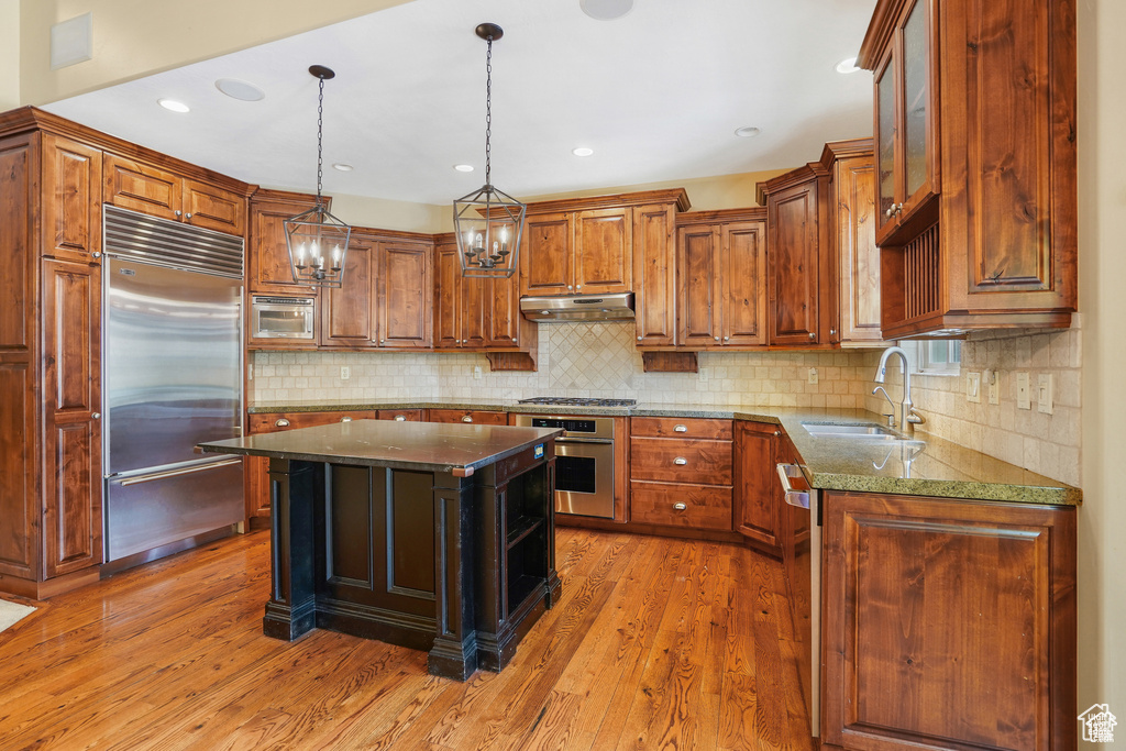 Kitchen with built in appliances, under cabinet range hood, light wood-type flooring, dark stone countertops, and hanging light fixtures