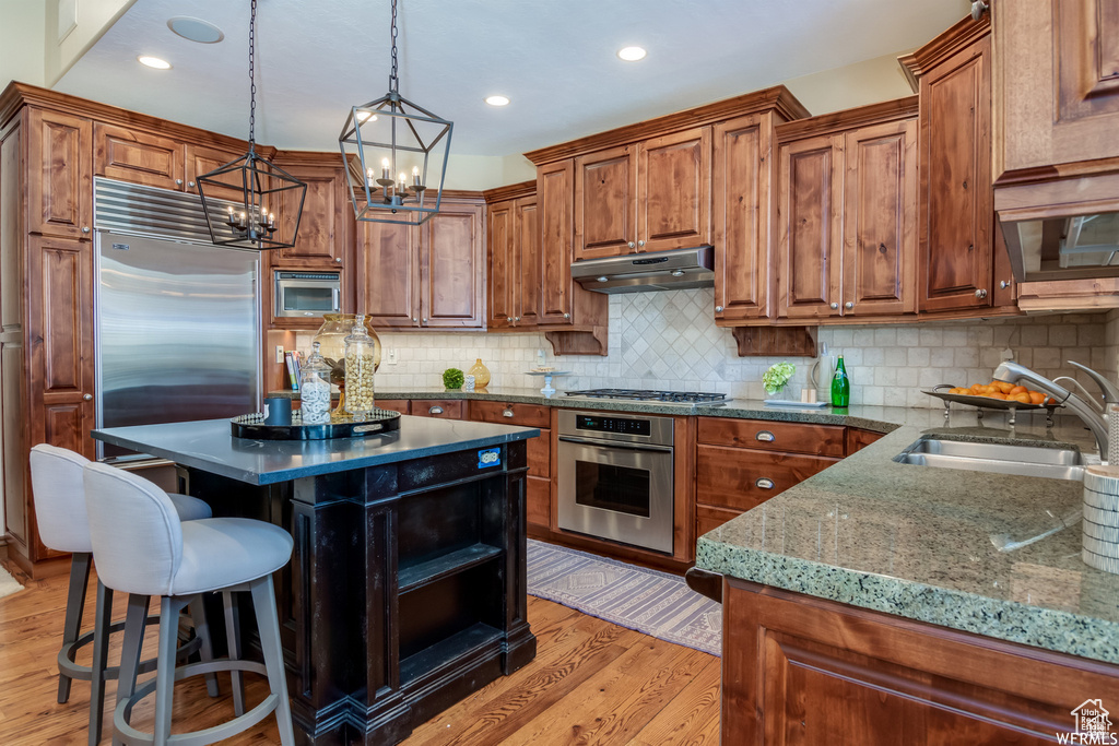Kitchen with built in appliances, under cabinet range hood, brown cabinets, a chandelier, and light wood-style flooring
