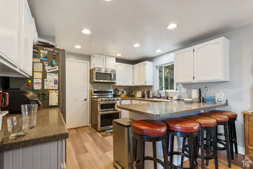 Kitchen with appliances with stainless steel finishes, a peninsula, white cabinets, light wood-style flooring, and recessed lighting