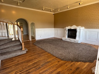 Unfurnished living room featuring rail lighting, ornamental molding, arched walkways, a wainscoted wall, and stairway