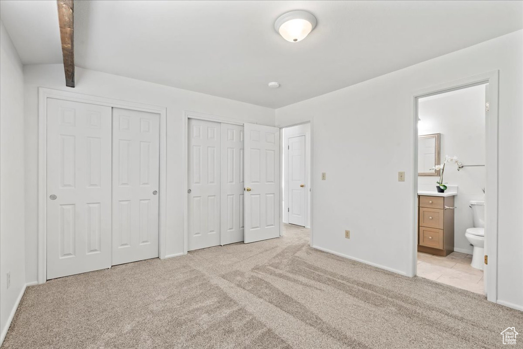 Unfurnished bedroom with two closets, light colored carpet, connected bathroom, and beamed ceiling