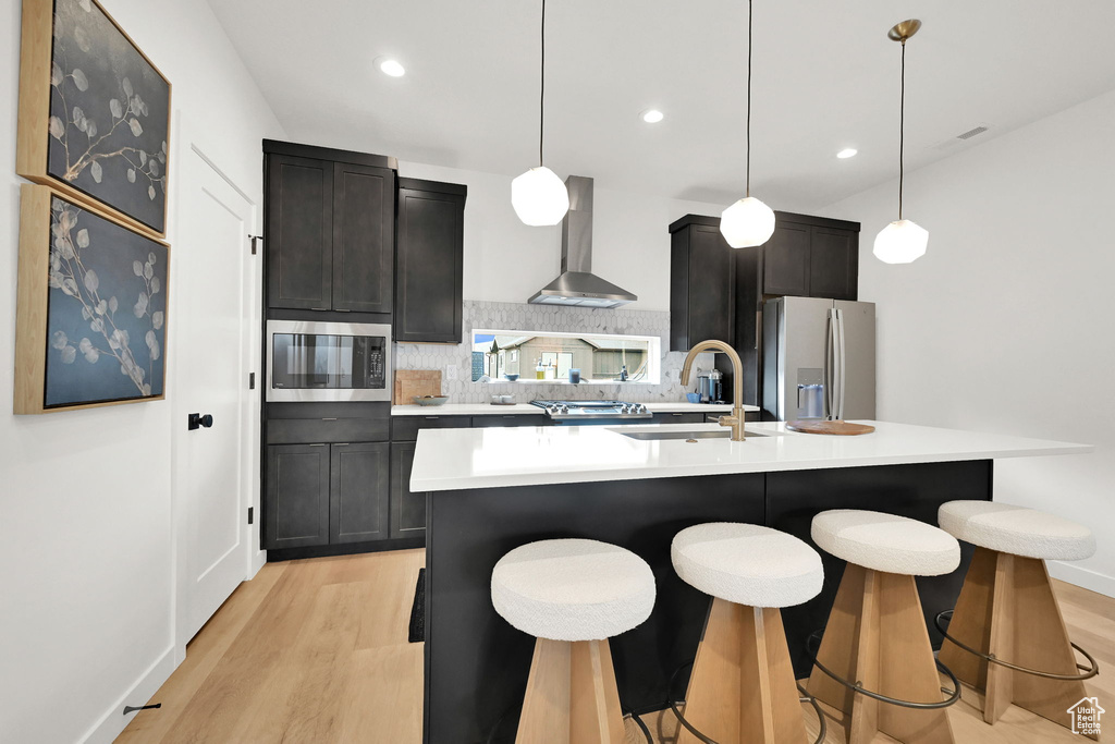 Kitchen with stainless steel appliances, wall chimney exhaust hood, backsplash, light wood-style floors, and a kitchen island with sink