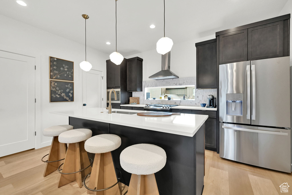 Kitchen with stainless steel fridge, light wood finished floors, wall chimney exhaust hood, light countertops, and a center island with sink