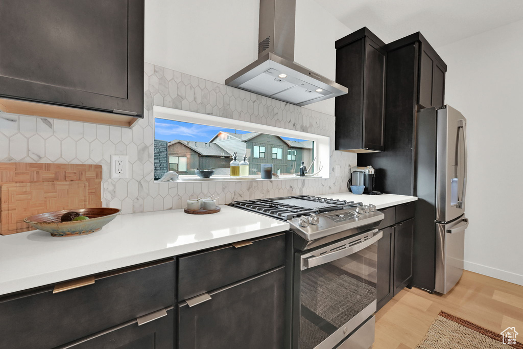 Kitchen featuring stainless steel appliances, wall chimney exhaust hood, light wood-style flooring, tasteful backsplash, and light countertops