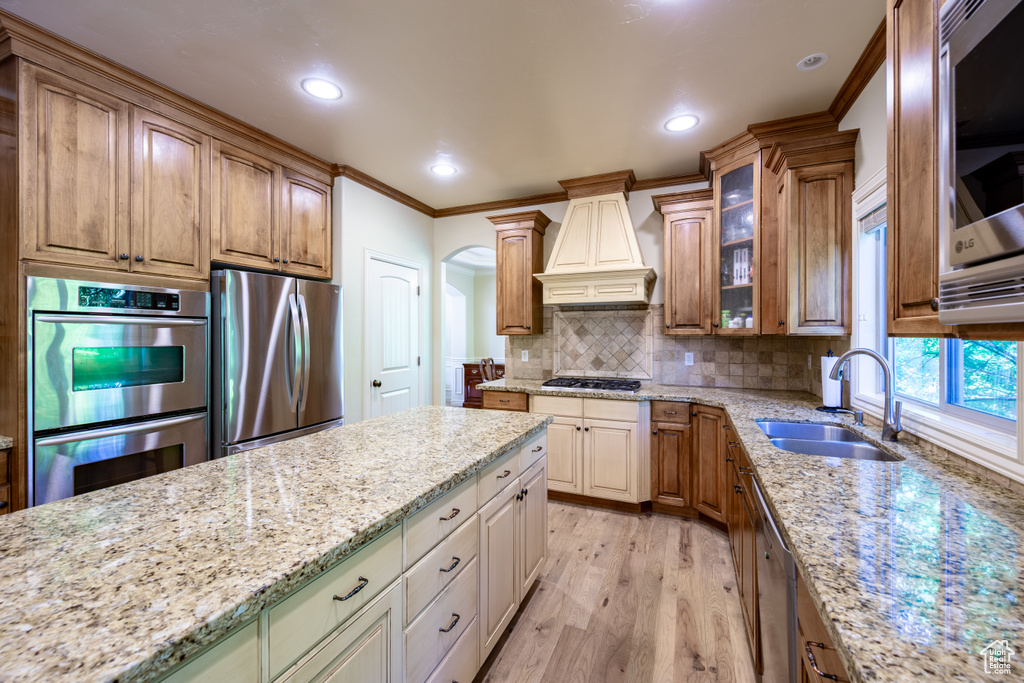 Kitchen featuring stainless steel appliances, custom range hood, crown molding, light stone countertops, and light wood-style floors