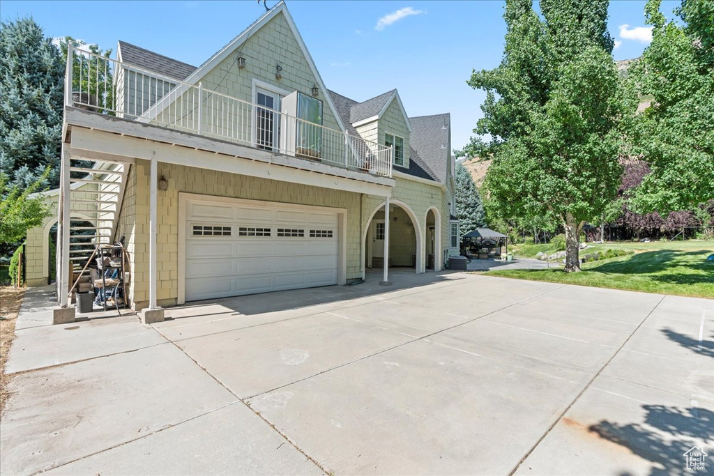 View of side of property with a balcony, concrete driveway, a shingled roof, an attached garage, and a yard