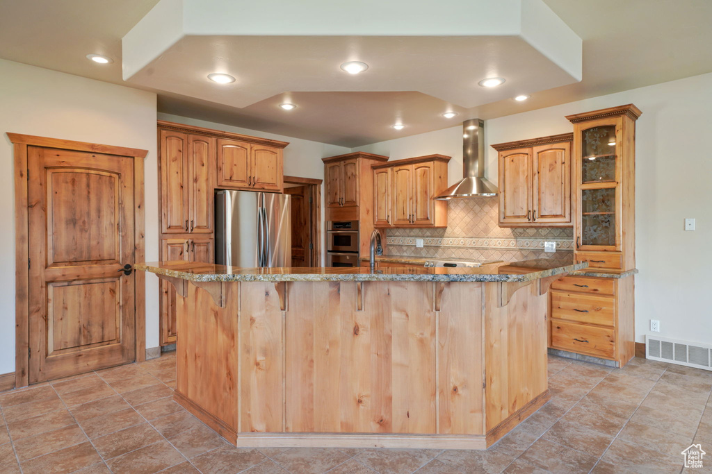 Kitchen featuring appliances with stainless steel finishes, wall chimney range hood, a breakfast bar, tasteful backsplash, and an island with sink