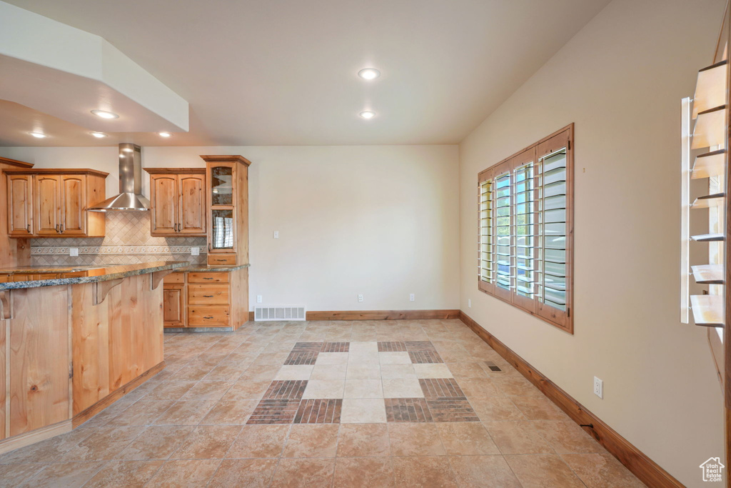 Kitchen featuring wall chimney range hood, a kitchen breakfast bar, decorative backsplash, glass insert cabinets, and recessed lighting