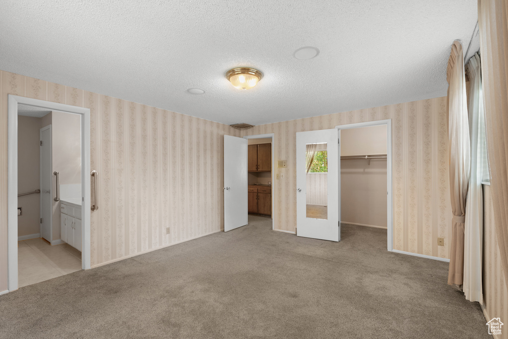 Unfurnished bedroom with wallpapered walls, a textured ceiling, carpet, a walk in closet, and ensuite bath