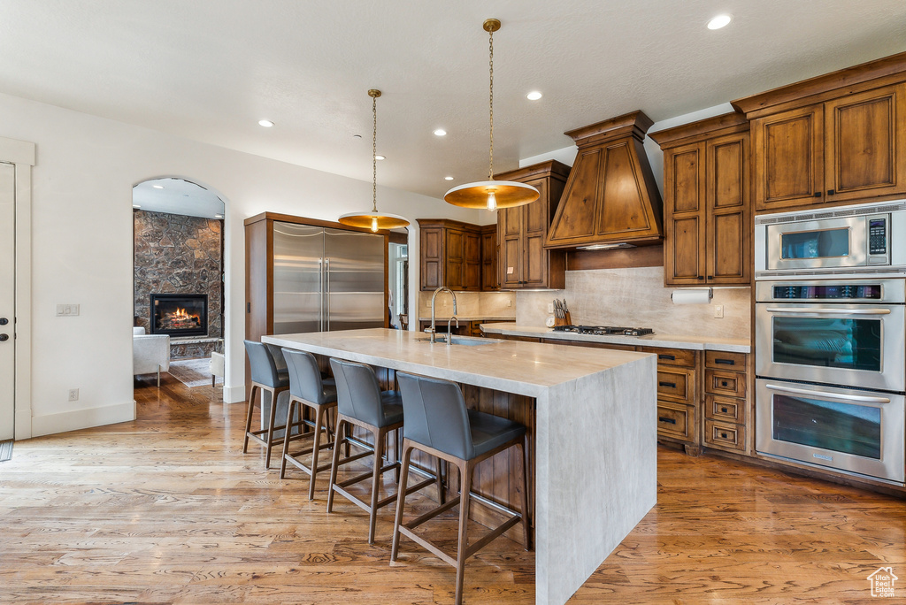 Kitchen with built in appliances, custom exhaust hood, a center island with sink, brown cabinets, and recessed lighting