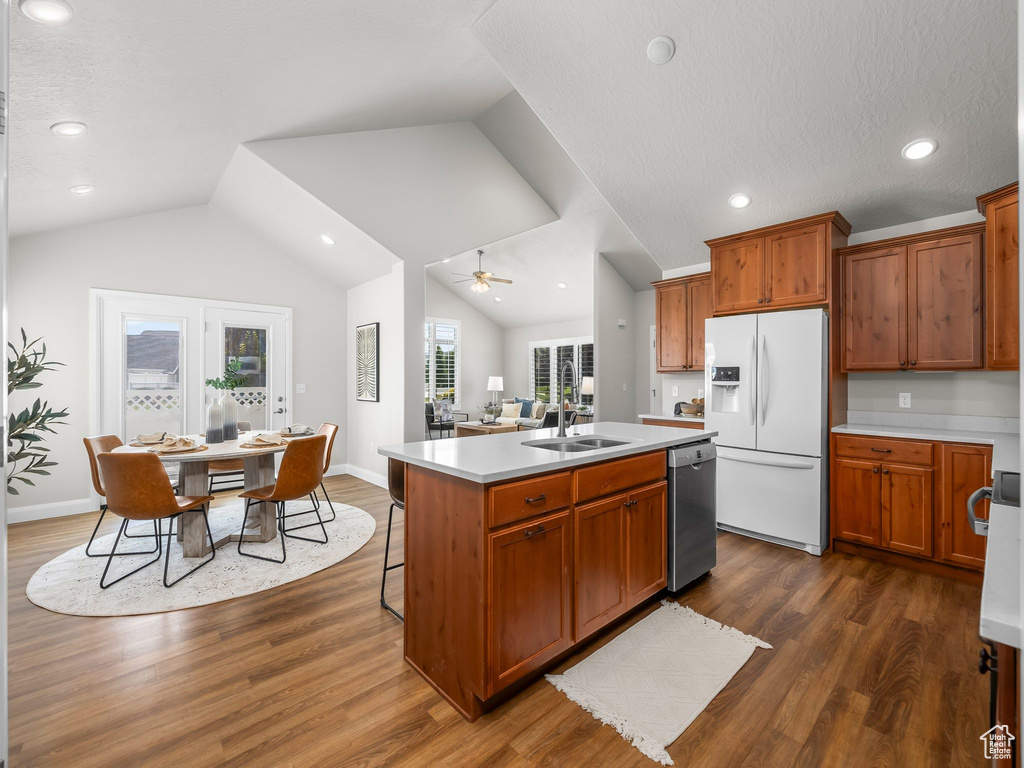 Kitchen featuring white refrigerator with ice dispenser, dishwasher, lofted ceiling, a center island with sink, and light countertops