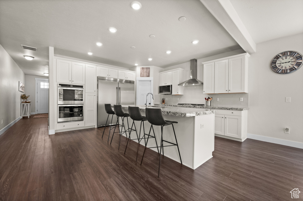 Kitchen featuring stainless steel appliances, wall chimney exhaust hood, a center island with sink, a kitchen breakfast bar, and white cabinetry