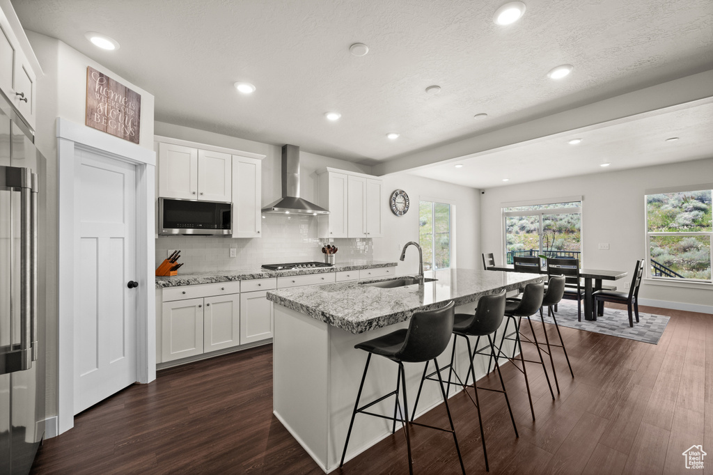 Kitchen with wall chimney exhaust hood, stainless steel appliances, dark wood finished floors, a breakfast bar, and a center island with sink