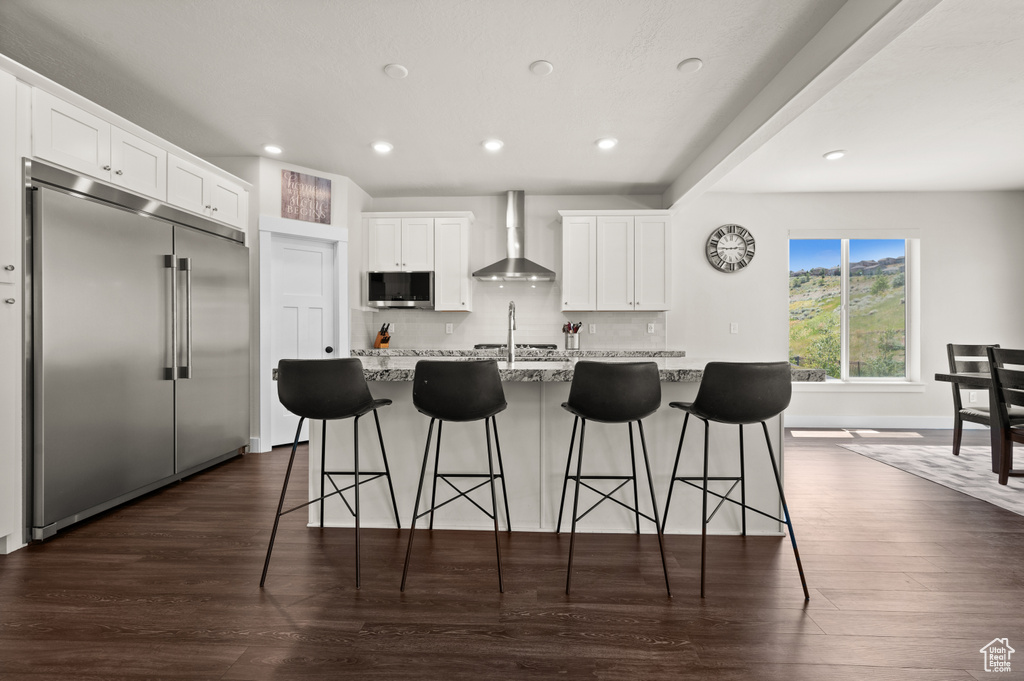 Kitchen featuring appliances with stainless steel finishes, wall chimney exhaust hood, an island with sink, dark wood finished floors, and white cabinets