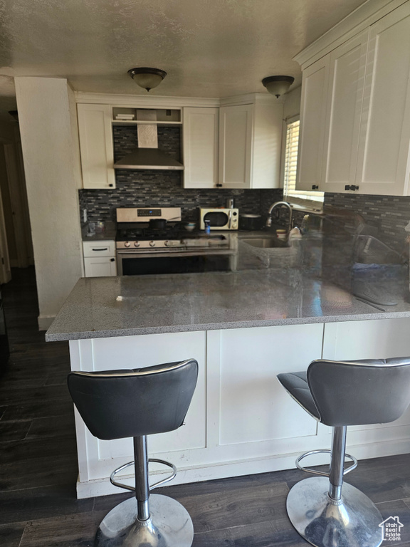 Kitchen featuring wall chimney range hood, stainless steel gas stove, a peninsula, white cabinetry, and tasteful backsplash