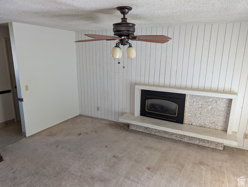 Unfurnished living room with ceiling fan, carpet flooring, a fireplace with raised hearth, wood walls, and a textured ceiling
