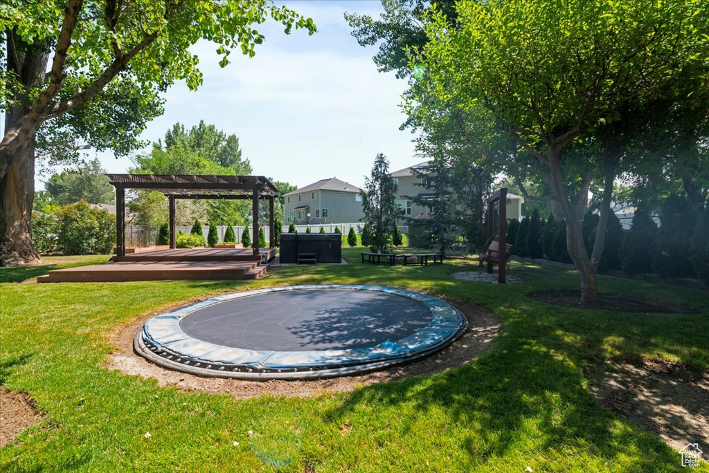 View of grassy yard featuring a pergola, a trampoline, a covered hot tub, and a deck