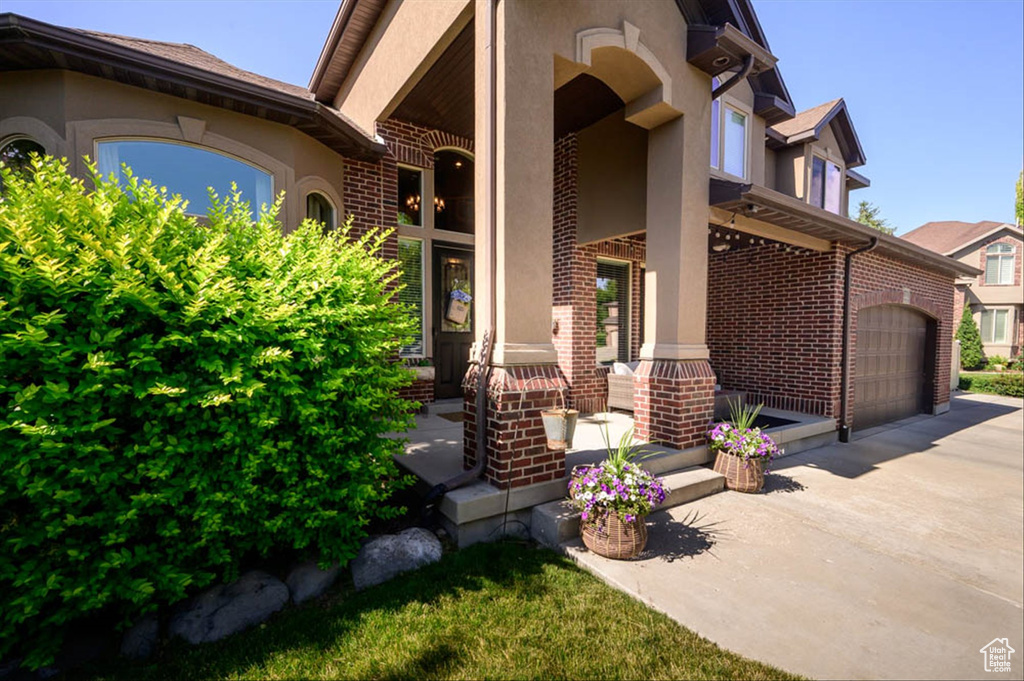 Doorway to property featuring stucco siding, covered porch, brick siding, an attached garage, and driveway