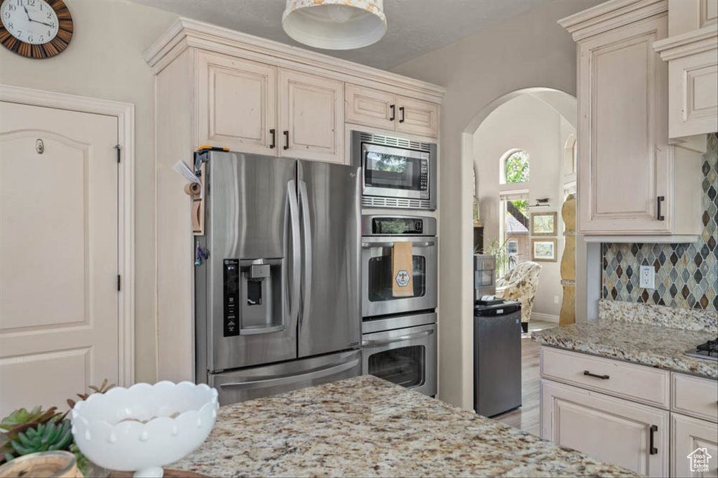 Kitchen featuring stainless steel appliances, arched walkways, tasteful backsplash, light stone countertops, and light wood finished floors