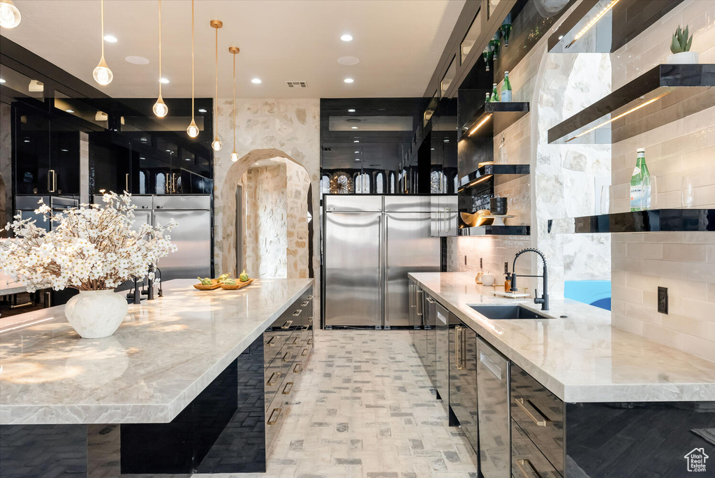 Kitchen featuring open shelves, dark cabinetry, built in fridge, light stone countertops, and decorative light fixtures