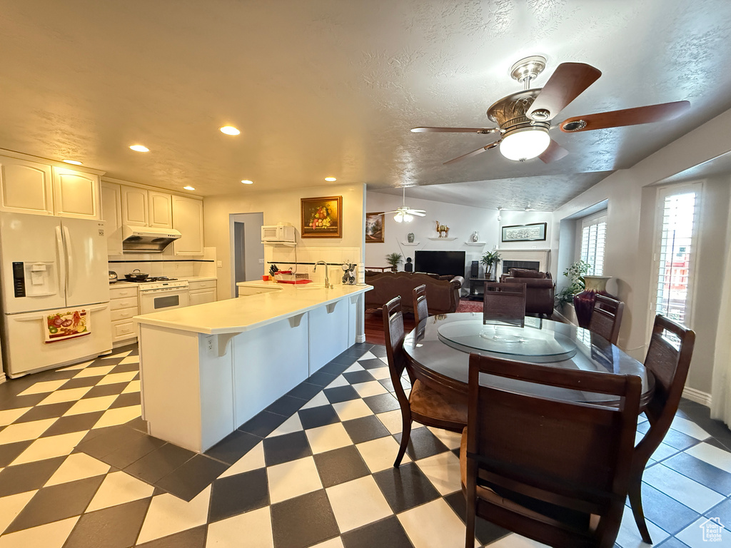 Kitchen with white appliances, a peninsula, a ceiling fan, a fireplace, and under cabinet range hood