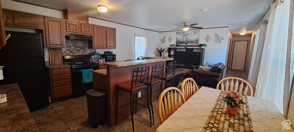 Kitchen featuring black appliances, under cabinet range hood, a kitchen bar, ceiling fan, and dark tile patterned flooring