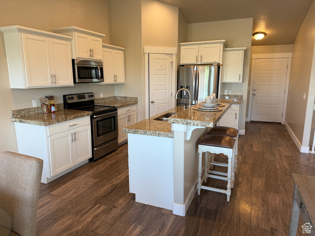 Kitchen featuring appliances with stainless steel finishes, dark wood-style flooring, white cabinets, light stone countertops, and a center island with sink