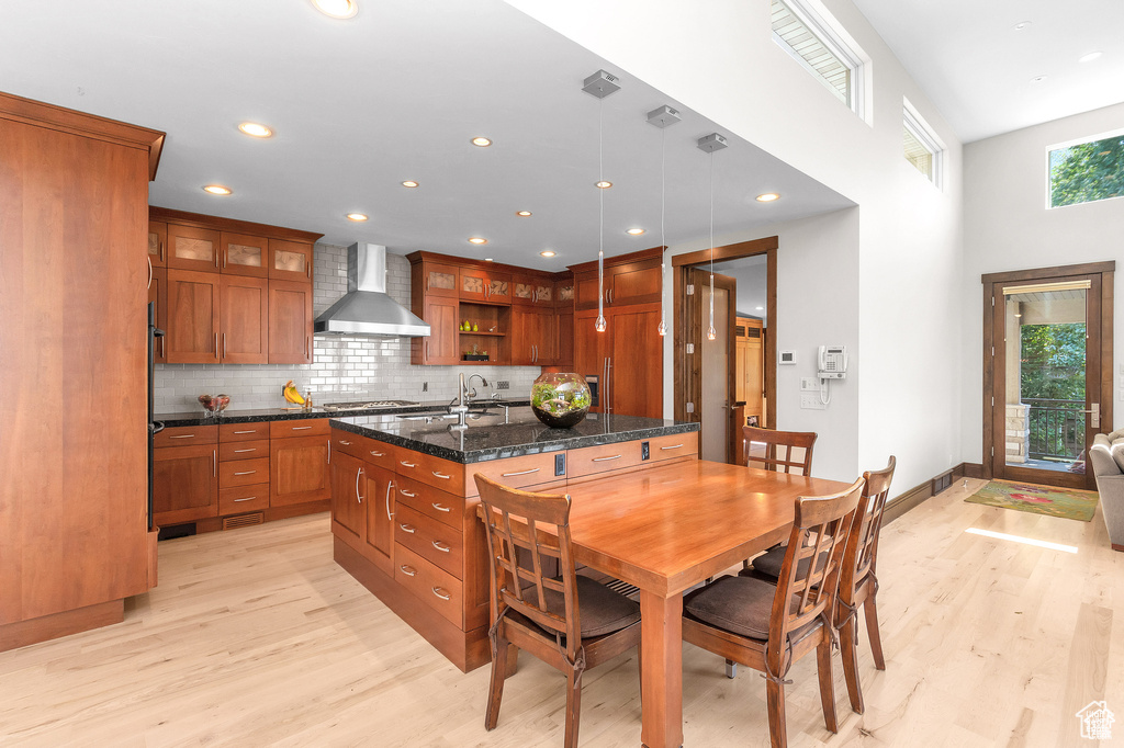 Kitchen featuring wall chimney range hood, brown cabinetry, stainless steel gas stovetop, light wood-style flooring, and glass insert cabinets