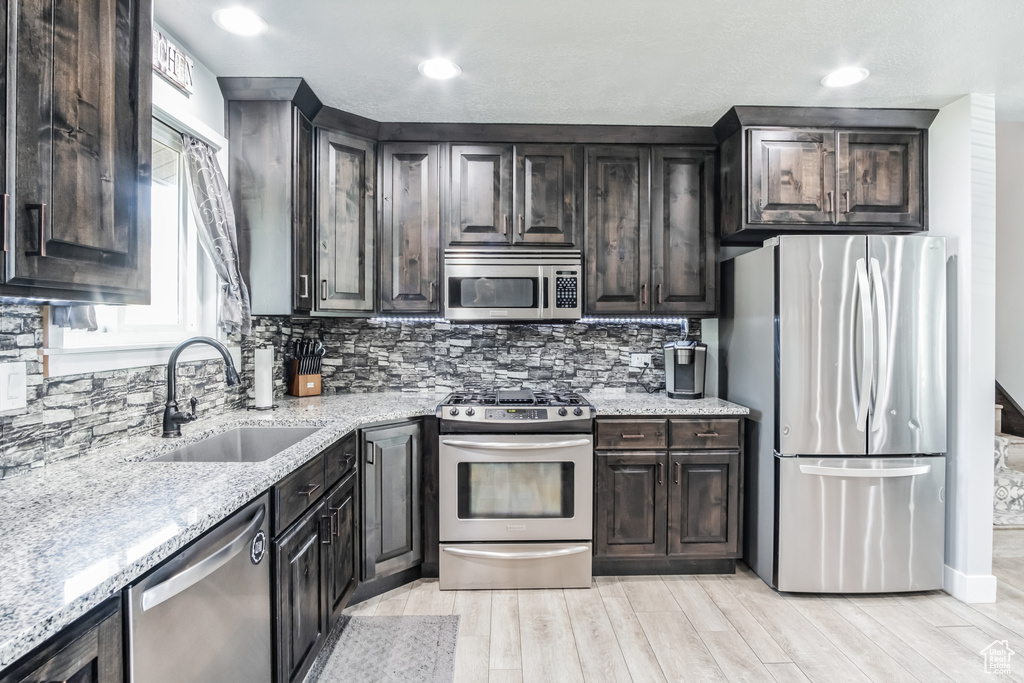 Kitchen with appliances with stainless steel finishes, light stone countertops, light wood-style flooring, decorative backsplash, and dark brown cabinetry