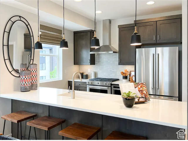 Kitchen featuring appliances with stainless steel finishes, wall chimney range hood, decorative backsplash, a kitchen breakfast bar, and pendant lighting
