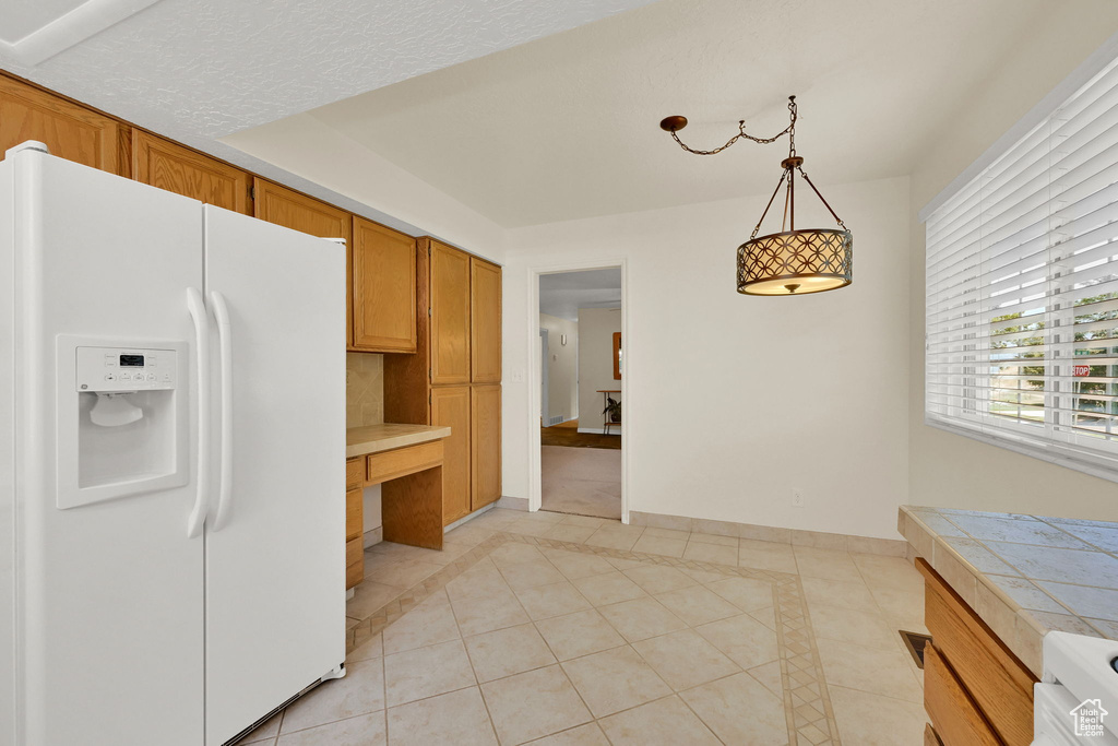 Kitchen featuring white refrigerator with ice dispenser, light tile patterned floors, hanging light fixtures, brown cabinets, and tile countertops