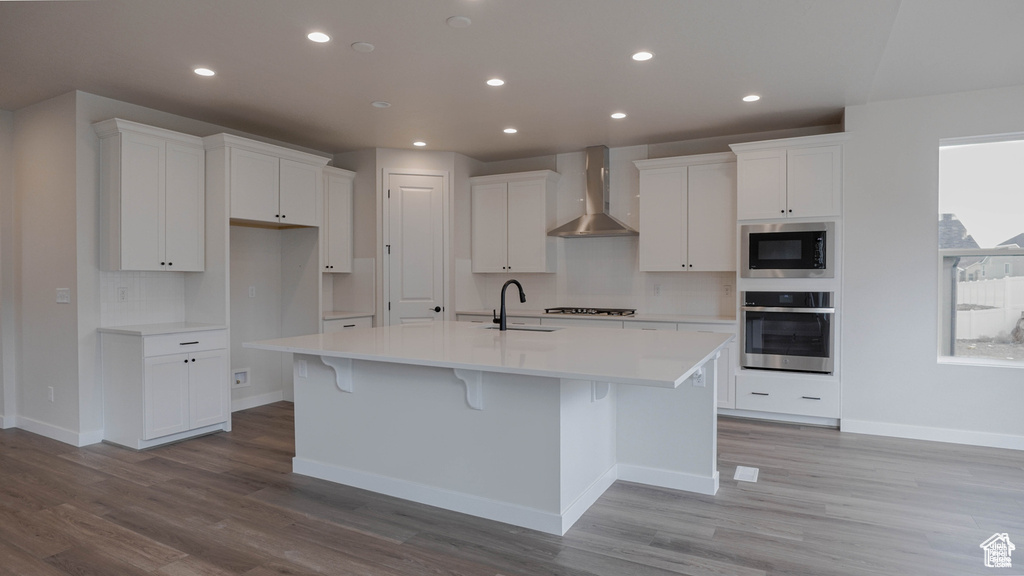 Kitchen with stainless steel appliances, wall chimney range hood, white cabinetry, light wood-style floors, and a spacious island