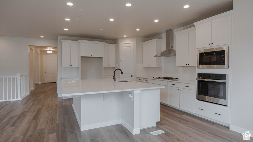 Kitchen featuring stainless steel appliances, wall chimney exhaust hood, a kitchen island with sink, light wood finished floors, and recessed lighting