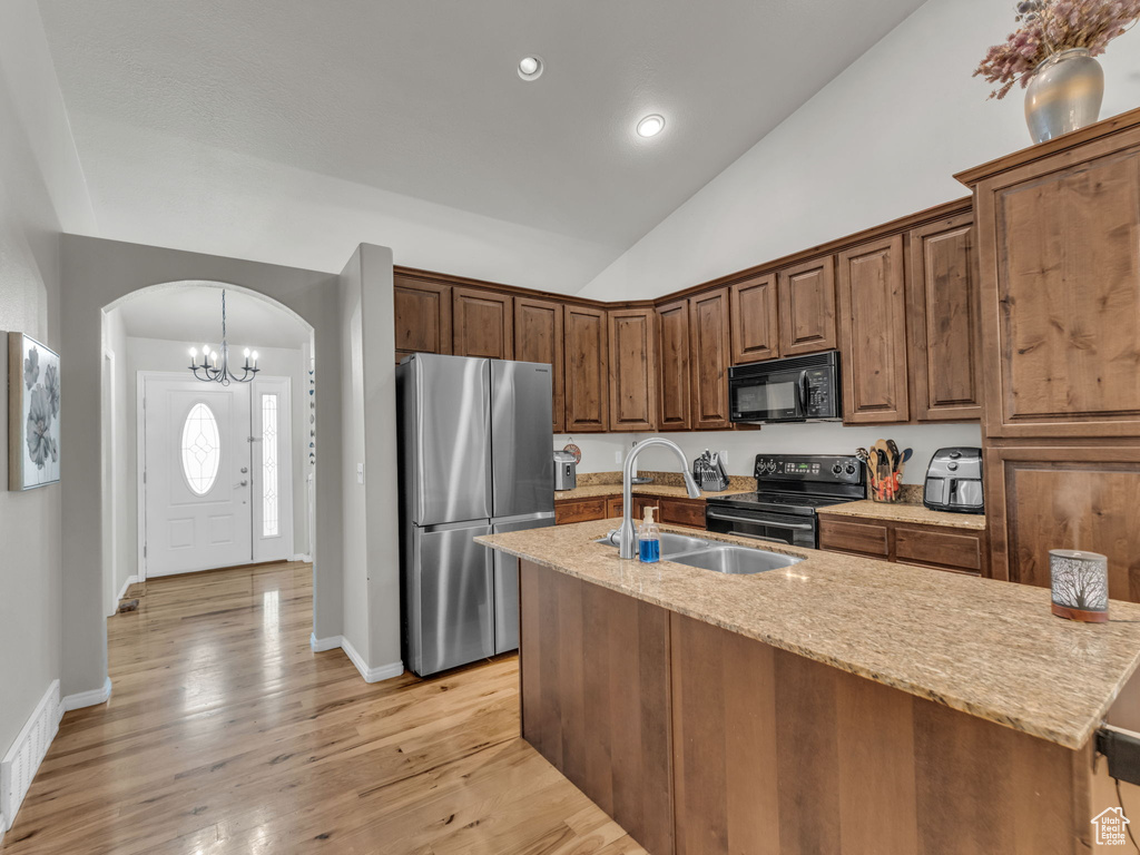 Kitchen featuring black appliances, arched walkways, a center island with sink, lofted ceiling, and light stone counters