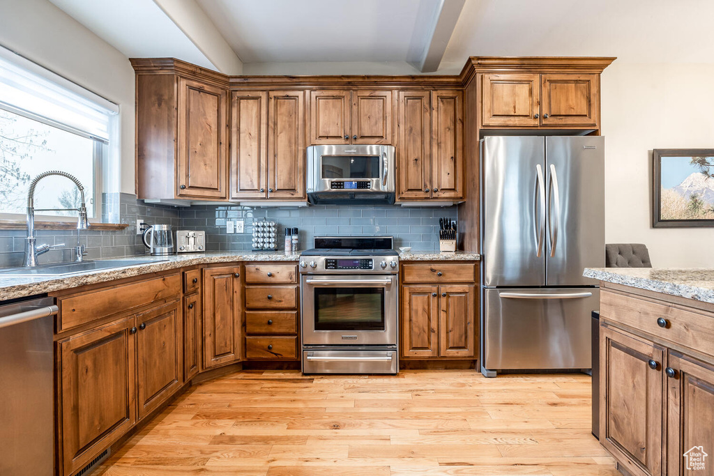 Kitchen with stainless steel appliances, brown cabinetry, light wood-type flooring, backsplash, and light stone counters