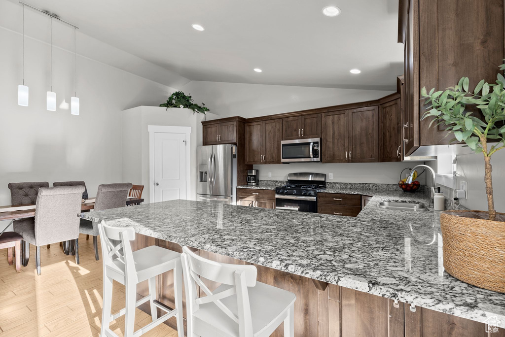 Kitchen with appliances with stainless steel finishes, lofted ceiling, light stone countertops, a peninsula, and dark brown cabinetry