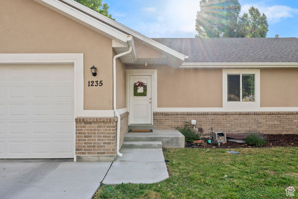 Doorway to property with brick siding, stucco siding, a yard, an attached garage, and roof with shingles