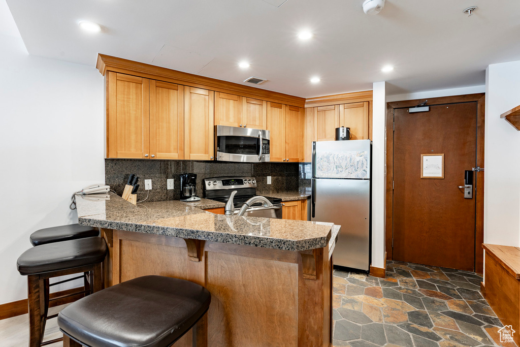 Kitchen featuring appliances with stainless steel finishes, a peninsula, decorative backsplash, a kitchen bar, and dark stone finish flooring