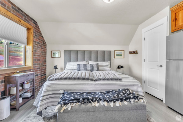 Bedroom with freestanding refrigerator, vaulted ceiling, light wood-style floors, and brick wall