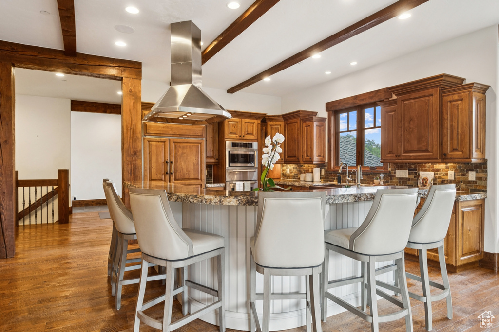 Kitchen with island exhaust hood, beam ceiling, backsplash, stainless steel oven, and light wood-style floors